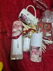 A set of items displayed on a red textured background including a decorative mug with floral and face designs, a bottle of scented home reed diffuser labeled 'Peony', and packaging with pink rose images. In the background, there are small wrapped gifts and what appears to be decorative red and white clips.
