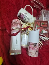 A set of items displayed on a red textured background including a decorative mug with floral and face designs, a bottle of scented home reed diffuser labeled 'Peony', and packaging with pink rose images. In the background, there are small wrapped gifts and what appears to be decorative red and white clips.