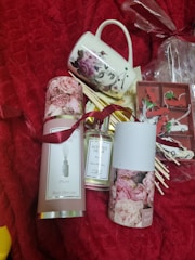 A set of items displayed on a red textured background including a decorative mug with floral and face designs, a bottle of scented home reed diffuser labeled 'Peony', and packaging with pink rose images. In the background, there are small wrapped gifts and what appears to be decorative red and white clips.