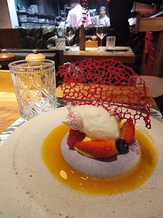 A gourmet dessert is elegantly presented on a round plate with a light textured surface. The dish features a dollop of cream atop a bed of sliced strawberries and a blueberry, surrounded by a golden sauce. A delicate red decorative lace piece adds a sophisticated touch. In the background, chefs in white uniforms are visible, working in an open kitchen.