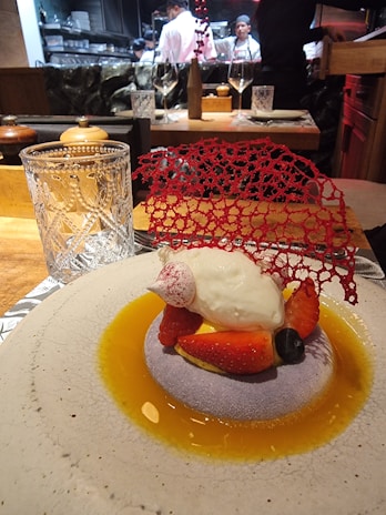 A gourmet dessert is elegantly presented on a round plate with a light textured surface. The dish features a dollop of cream atop a bed of sliced strawberries and a blueberry, surrounded by a golden sauce. A delicate red decorative lace piece adds a sophisticated touch. In the background, chefs in white uniforms are visible, working in an open kitchen.