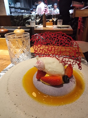 A gourmet dessert is elegantly presented on a round plate with a light textured surface. The dish features a dollop of cream atop a bed of sliced strawberries and a blueberry, surrounded by a golden sauce. A delicate red decorative lace piece adds a sophisticated touch. In the background, chefs in white uniforms are visible, working in an open kitchen.