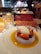 A gourmet dessert is elegantly presented on a round plate with a light textured surface. The dish features a dollop of cream atop a bed of sliced strawberries and a blueberry, surrounded by a golden sauce. A delicate red decorative lace piece adds a sophisticated touch. In the background, chefs in white uniforms are visible, working in an open kitchen.