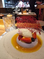 A gourmet dessert is elegantly presented on a round plate with a light textured surface. The dish features a dollop of cream atop a bed of sliced strawberries and a blueberry, surrounded by a golden sauce. A delicate red decorative lace piece adds a sophisticated touch. In the background, chefs in white uniforms are visible, working in an open kitchen.