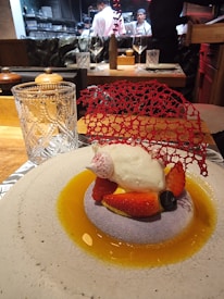 A gourmet dessert is elegantly presented on a round plate with a light textured surface. The dish features a dollop of cream atop a bed of sliced strawberries and a blueberry, surrounded by a golden sauce. A delicate red decorative lace piece adds a sophisticated touch. In the background, chefs in white uniforms are visible, working in an open kitchen.