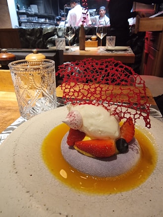 A gourmet dessert is elegantly presented on a round plate with a light textured surface. The dish features a dollop of cream atop a bed of sliced strawberries and a blueberry, surrounded by a golden sauce. A delicate red decorative lace piece adds a sophisticated touch. In the background, chefs in white uniforms are visible, working in an open kitchen.