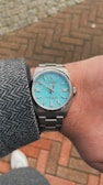 A luxury wristwatch with a silver metal band and a turquoise-colored face featuring silver markers and hands. It is worn on a person's wrist, covered by a gray tweed jacket sleeve. The background consists of a red and beige brick pavement.