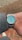 A luxury wristwatch with a silver metal band and a turquoise-colored face featuring silver markers and hands. It is worn on a person's wrist, covered by a gray tweed jacket sleeve. The background consists of a red and beige brick pavement.