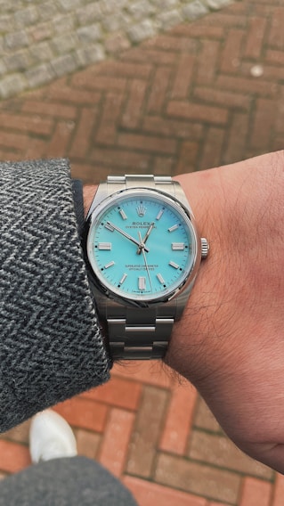 A luxury wristwatch with a silver metal band and a turquoise-colored face featuring silver markers and hands. It is worn on a person's wrist, covered by a gray tweed jacket sleeve. The background consists of a red and beige brick pavement.