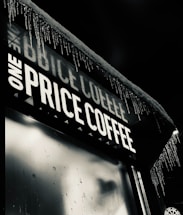 An outdoor signboard with the words 'ONE PRICE COFFEE' illuminated in bright white light. Icicles are hanging from the edge of the roof, indicating cold weather conditions. The background is dark, suggesting that the scene is happening at night.