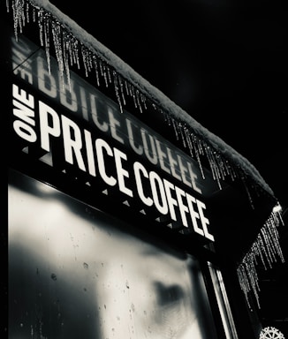 An outdoor signboard with the words 'ONE PRICE COFFEE' illuminated in bright white light. Icicles are hanging from the edge of the roof, indicating cold weather conditions. The background is dark, suggesting that the scene is happening at night.