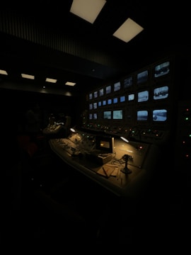 A dimly lit control room with a panel displaying multiple monitors showing various black-and-white images. The room features several control panels with switches, buttons, and indicator lights. A desk lamp illuminates part of the workspace, where papers, a microphone, and some equipment are placed neatly.