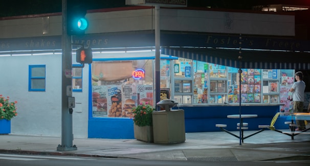 A fast-food restaurant, featuring a retro-style exterior, has an illuminated 'open' sign and windows showcasing menu items. A lone person stands near tables outside, possibly waiting for an order. Potted plants with flowers adorn the sidewalk, and a traffic light with a pedestrian signal is in view.