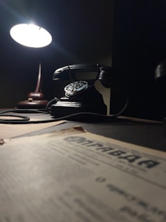 A dimly lit scene featuring a vintage rotary phone on a wooden desk. A bright desk lamp casts a soft glow over the phone, creating a nostalgic atmosphere. In the foreground, there is a blurred newspaper or document with typography, adding to the antique feel.