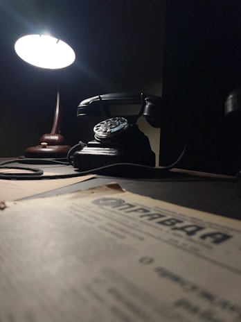 A dimly lit scene featuring a vintage rotary phone on a wooden desk. A bright desk lamp casts a soft glow over the phone, creating a nostalgic atmosphere. In the foreground, there is a blurred newspaper or document with typography, adding to the antique feel.