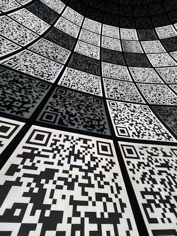 A geometric arrangement of numerous QR codes fills the perspective with a grid-like pattern. The codes are aligned in a curved fashion, creating a sense of depth and a digital, futuristic atmosphere.