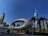 A futuristic building with an elliptical structure covered in Arabic calligraphy stands prominently against a clear blue sky. Skyscrapers and palm trees surround the area, contributing to a modern urban landscape. People walk alongside a road, and a red vehicle is visible nearby.