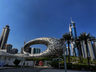 A futuristic building with an elliptical structure covered in Arabic calligraphy stands prominently against a clear blue sky. Skyscrapers and palm trees surround the area, contributing to a modern urban landscape. People walk alongside a road, and a red vehicle is visible nearby.