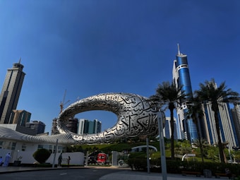 A futuristic building with an elliptical structure covered in Arabic calligraphy stands prominently against a clear blue sky. Skyscrapers and palm trees surround the area, contributing to a modern urban landscape. People walk alongside a road, and a red vehicle is visible nearby.