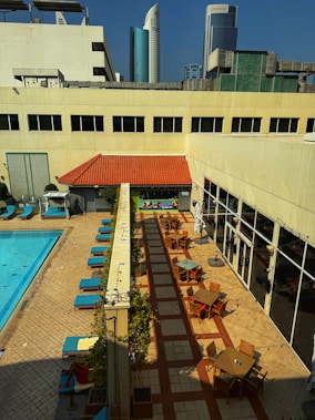 An outdoor area with a swimming pool lined with blue lounge chairs. Nearby, there is a patio with wooden tables and chairs under umbrellas. The architecture is modern, and several high-rise buildings are visible in the background under a clear blue sky.