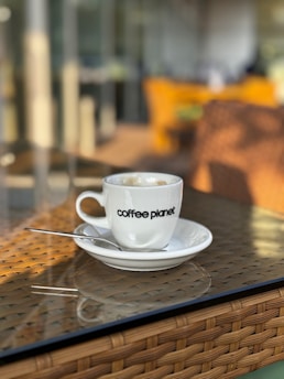 A coffee cup with the words 'coffee planet' on it sits on a saucer with a spoon, placed on a woven rattan table with a glass top. The background is blurred, showcasing a warm, inviting indoor or outdoor cafe setting.