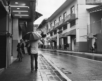 A black and white street scene captured with a Leica, showing a lone figure walking under a rain-soaked umbrella.
