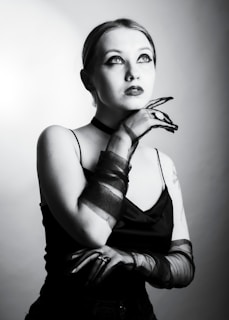 A woman gazes upward with a thoughtful expression, wearing dark makeup and sheer gloves. Her hand is elegantly positioned near her chin, and she is dressed in a sleeveless black top. The lighting creates a dramatic, high-contrast effect in black and white.