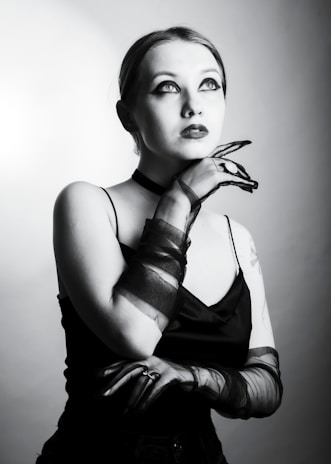 A woman gazes upward with a thoughtful expression, wearing dark makeup and sheer gloves. Her hand is elegantly positioned near her chin, and she is dressed in a sleeveless black top. The lighting creates a dramatic, high-contrast effect in black and white.