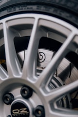 A close-up view of a vehicle wheel featuring a silver alloy rim with a sporty design. The rim has multiple spokes and displays the manufacturer's logo at the center. Behind the rim, a ventilated disc brake is visible with a recognizable emblem.