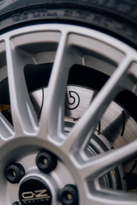 A close-up view of a vehicle wheel featuring a silver alloy rim with a sporty design. The rim has multiple spokes and displays the manufacturer's logo at the center. Behind the rim, a ventilated disc brake is visible with a recognizable emblem.