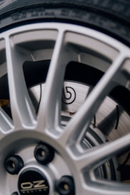 A close-up view of a vehicle wheel featuring a silver alloy rim with a sporty design. The rim has multiple spokes and displays the manufacturer's logo at the center. Behind the rim, a ventilated disc brake is visible with a recognizable emblem.