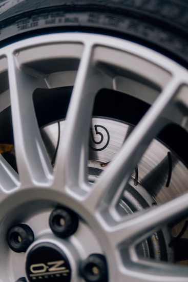 A close-up view of a vehicle wheel featuring a silver alloy rim with a sporty design. The rim has multiple spokes and displays the manufacturer's logo at the center. Behind the rim, a ventilated disc brake is visible with a recognizable emblem.