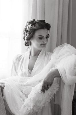 A woman with styled hair is wearing a robe and holding a delicate lace garment. She is looking down at the garment with a gentle smile, standing near a window with soft light illuminating her face.