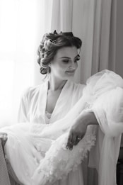 A woman with styled hair is wearing a robe and holding a delicate lace garment. She is looking down at the garment with a gentle smile, standing near a window with soft light illuminating her face.