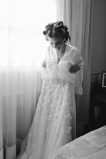 A person stands in front of a window with delicate sheer curtains, holding up a detailed bridal gown adorned with floral appliques. The individual has hair styled in rollers and is wearing a robe. The setting is a cozy room with soft lighting.