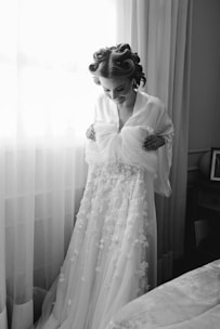 A person stands in front of a window with delicate sheer curtains, holding up a detailed bridal gown adorned with floral appliques. The individual has hair styled in rollers and is wearing a robe. The setting is a cozy room with soft lighting.