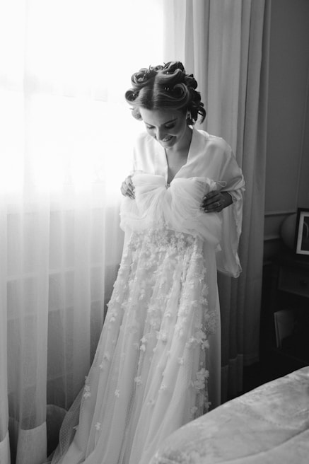 A person stands in front of a window with delicate sheer curtains, holding up a detailed bridal gown adorned with floral appliques. The individual has hair styled in rollers and is wearing a robe. The setting is a cozy room with soft lighting.