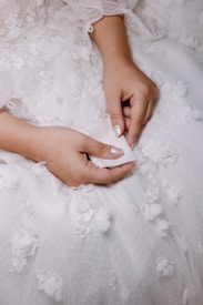 Delicate hands resting on a white, intricately embroidered dress with floral designs, holding a white fabric.
