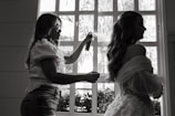 A stylist is preparing a woman for an event, possibly a wedding, by applying hairspray to her hairstyle. The setting is indoors with natural light coming through a window, casting silhouettes of both individuals. The woman wears an elaborate gown featuring lace and floral details.