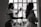 A stylist is preparing a woman for an event, possibly a wedding, by applying hairspray to her hairstyle. The setting is indoors with natural light coming through a window, casting silhouettes of both individuals. The woman wears an elaborate gown featuring lace and floral details.