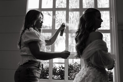 A stylist is preparing a woman for an event, possibly a wedding, by applying hairspray to her hairstyle. The setting is indoors with natural light coming through a window, casting silhouettes of both individuals. The woman wears an elaborate gown featuring lace and floral details.