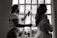 A stylist is preparing a woman for an event, possibly a wedding, by applying hairspray to her hairstyle. The setting is indoors with natural light coming through a window, casting silhouettes of both individuals. The woman wears an elaborate gown featuring lace and floral details.