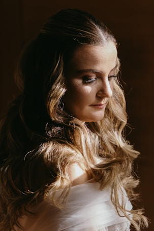 A woman with long, wavy hair cascading over her shoulders, gazing downwards. Soft lighting accentuates her delicate features and highlights the texture in her hair. The background is dark, adding to the focus on her serene expression.
