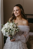 A bride is dressed in an elegant, white wedding gown with intricate lace and floral details. She is holding a bouquet of white flowers, and her hair is styled in loose waves with a natural makeup look. The setting is indoors, near a softly lit window, creating a serene and warm atmosphere.