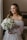 A bride is dressed in an elegant, white wedding gown with intricate lace and floral details. She is holding a bouquet of white flowers, and her hair is styled in loose waves with a natural makeup look. The setting is indoors, near a softly lit window, creating a serene and warm atmosphere.
