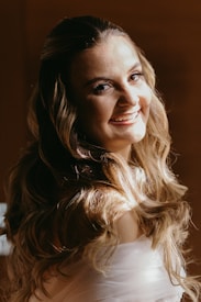A woman with long, wavy hair smiles warmly at the camera. The light highlights her hair and facial features, creating a soft glow. The background is a warm, muted brown, adding to the intimate and cozy atmosphere.