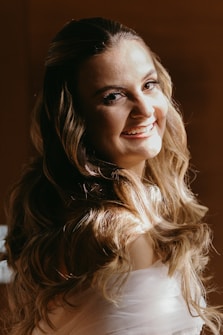 A woman with long, wavy hair smiles warmly at the camera. The light highlights her hair and facial features, creating a soft glow. The background is a warm, muted brown, adding to the intimate and cozy atmosphere.