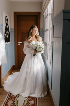A radiant bride in a flowing white gown stands against a soft baby blue backdrop, holding a delicate bouquet.