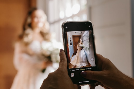 A person is holding a smartphone, capturing a photo of a bride dressed in a wedding gown. The bride stands near a window, holding a bouquet, with soft light illuminating her.