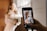 A person is holding a smartphone, capturing a photo of a bride dressed in a wedding gown. The bride stands near a window, holding a bouquet, with soft light illuminating her.